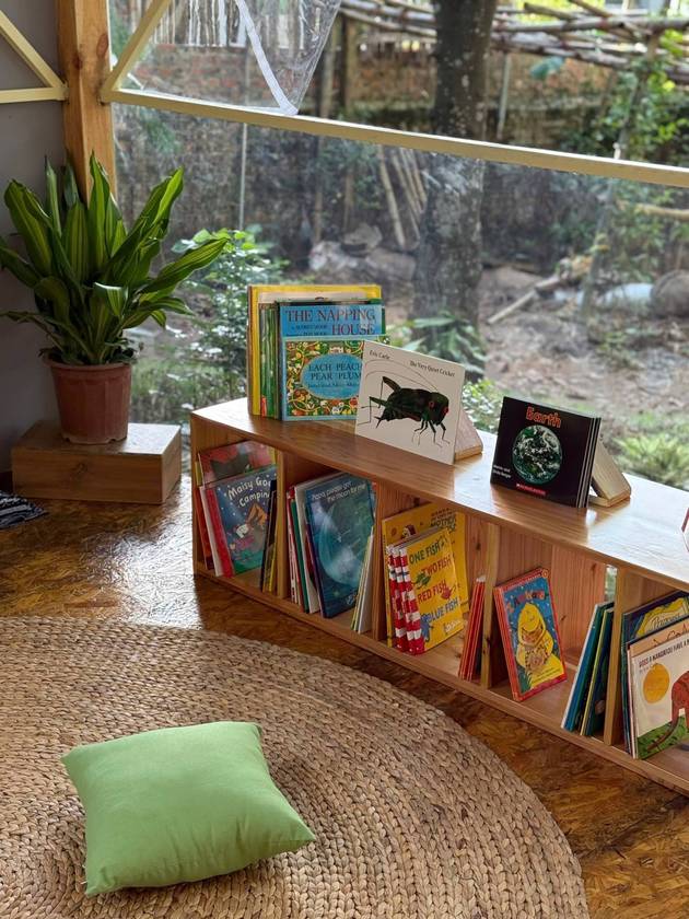 Low wooden bookshelf of picture books beside a woven rug and green cushion, looking out onto the garden